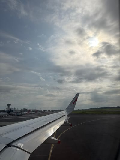 The image captures a detailed view from inside a commercial passenger aircraft on an airport tarmac in San Luis Talpa, El Salvador, specifically at Oscar Arnulfo Romero International Airport (MSLP).

In the foreground, the bright white wing of the aircraft from which the photograph is taken extends from the lower left to the mid-right of the frame. The wing displays a red, stylized bird or 'A' logo on its upturned winglet, consistent with Avianca livery. The underside of the wing shows visible flap tracks and structural elements.

The tarmac, dark grey asphalt with yellow painted lines, occupies the lower mid-ground. To the left, several other commercial airliners are parked at gates, some featuring red liveries. A prominent, multi-tiered air traffic control tower, predominantly white and grey, is visible beyond the parked aircraft and terminal buildings.

To the right, beyond the aircraft wing, the tarmac extends into a section of green grass or field, bordered by a distant line of trees on the horizon. The sky is largely overcast with extensive grey clouds. A bright, diffused light source, indicating the sun, is visible through a break in the clouds in the upper right quadrant, casting a strong glare. No individual people or explicit interactions are discernible within the frame. The scene suggests the aircraft is either stationary or slowly taxiing on the airport surface under cloudy conditions.