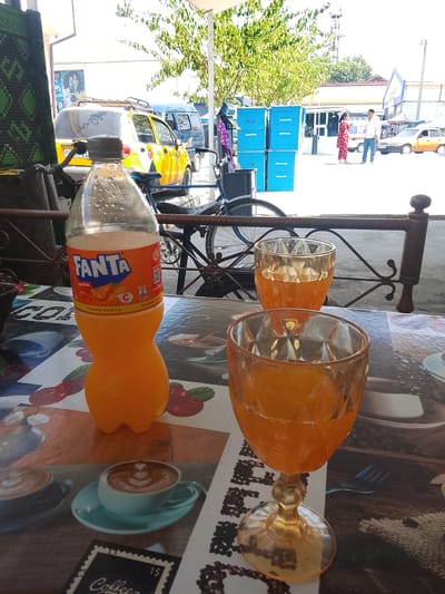 In the city of Sariosiyo, Uzbekistan, an outdoor table is set with a bottle of Fanta orange soda and two glasses of orange beverage. The table is covered with a printed cloth depicting coffee cups and fruit. In the background, a yellow car is parked, and several blue storage units are stacked. Two individuals, a woman in a red patterned dress and a man in a white shirt and blue pants, are standing near the storage units. The scene is illuminated by daylight.