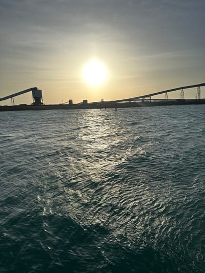 The image depicts a body of water with gentle waves, reflecting the sun's bright glare. In the background, a long industrial structure stretches across the horizon, characterized by conveyor belts and silos, likely part of a port or mining facility. The sky above is a gradient of soft yellows and grays, with the sun positioned prominently in the upper center, partially obscured by a light haze. The overall scene suggests an industrial waterfront during a sunset or sunrise. This location is consistent with the port facilities in Uribia, Colombia, which handle bulk materials like coal and salt.
