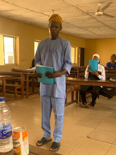 This image captures an indoor classroom setting within an educational facility in Abobo, Nigeria.
In the foreground, a young male subject stands centrally, facing slightly right. He wears a light blue traditional Nigerian tunic and trousers, a striped yellow and brown headwrap (gẹlẹ), and black slide sandals. He holds a light blue folder containing white papers in both hands at chest level, looking forward with a neutral expression.
To the right of the central subject, in the midground, a female student is seated at a wooden desk. She wears a white head covering, a white long-sleeved top, and black trousers with white sandals. She holds a light blue folder directly in front of her face, obscuring it. A black bag with a white "CHANEL" logo is visible on the floor beside her desk. Further back and to the right, another male student is seated, wearing a patterned cap and a blue shirt, appearing to observe something to his right.
The classroom features multiple rows of wooden desks and benches, typical of an academic environment. The floor is covered with light beige or tan square tiles. The walls are painted a light yellow or cream color. The ceiling is white with square acoustic panels, and a white four-blade ceiling fan, labeled "POWER METERS," is mounted near the center. Windows allowing natural light are visible on the left side of the room.
On a desk in the immediate lower-left foreground, a clear plastic bottle of CWAY water and a silver and orange aluminum can of Maltina are present.
