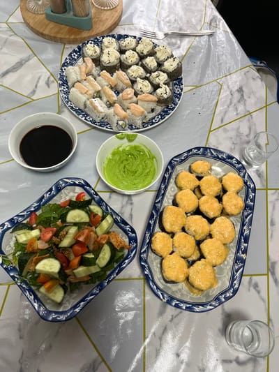 The image shows a table set with a meal, likely a dinner or lunch, in Октябрьское, Kazakhstan. The scene is set indoors, with a marble-patterned tablecloth covering the table.
The meal consists of several dishes. On a blue and white patterned oval platter are many pieces of sushi, topped with a creamy, orange sauce and sprinkled with black sesame seeds. To the left, another blue and white patterned plate holds more sushi, some topped with a white sauce and black sesame seeds, and others with pieces of raw salmon. In the center, a small white bowl contains a vibrant green sauce, possibly avocado-based. Next to it, a small white bowl holds dark soy sauce. To the right of the platter with the orange-sauced sushi, a long, rectangular blue and white patterned dish contains a salad. The salad is made with mixed greens, sliced cucumbers, halved cherry tomatoes, and pieces of grilled chicken.
There are also two glasses of water on the table. A fork is visible on the left side of the sushi plate. A wooden cutting board with a salt and pepper shaker set is partially visible on the far left.
The lighting suggests it could be daytime or evening, but there are no strong indicators of weather. The overall impression is a home-cooked meal, possibly for a family gathering or a casual dinner. There is no visible text in the image.