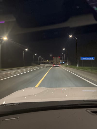 The image depicts a nighttime scene on a highway, likely in or near Таймурзино, Russia, based on the road sign indicating "ATCYPOBO 4" (likely referring to the direction of Ацырово, 4 kilometers away). The view is from inside a vehicle, looking forward. A truck is visible in the distance. Streetlights illuminate the road. Road markings include white lane dividers and a yellow line. The foreground shows part of the interior of the vehicle, specifically the dashboard and the car's body.