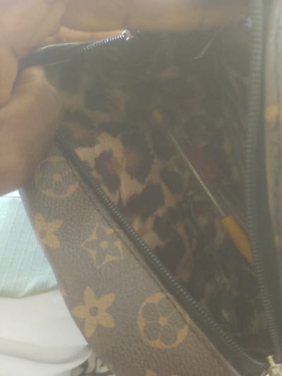 An interior view of an open brown designer bag with a leopard print lining is captured. The bag's exterior features the iconic LV monogram pattern. Two writing instruments, one with a red top and the other with a yellow top, are visible inside. The bag is resting on a white and light blue textured surface, possibly a bed or sofa. The image is a close-up shot, with a hand partially visible on the left side, holding the bag open. The scene is dimly lit and slightly out of focus. There is no indication of the city of Majemu, Nigeria, in the image or its context.