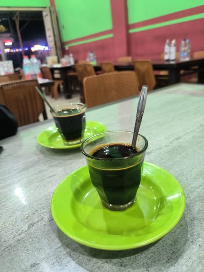 Two glasses of dark liquid, likely coffee, are served on bright green saucers on a light gray, speckled table. Each glass contains a spoon. The glass in the foreground is closer to the viewer and slightly out of focus, while the glass behind it is also out of focus. The background shows the interior of what appears to be a restaurant or cafe. Rows of tables and chairs, some made of wicker and others darker wood, are visible. The walls are painted with a green upper section and a reddish-pink lower section, separated by a horizontal stripe. Bottles of water are visible on a table in the background. The lighting suggests it is either evening or indoors with artificial lighting, as there are also some blurred colorful lights visible in the distance. The overall scene is casual and appears to be set in Meukokuthang, Indonesia, based on the location context. No text is visible in the image.