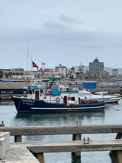 A medium shot captures a blue and white fishing boat, the "GUIMERA II PT 21142 EM," docked in a harbor. The boat, with its dark blue hull and white superstructure, has a small cabin with red doors and windows. Several life preservers are visible on its exterior. Two flags fly from the mast, one with a red, blue, and yellow design, and another with a red and white checkered pattern. In the background, a cityscape unfolds, featuring various buildings of different heights and architectural styles. Prominent among them is a tall, modern building with a blue facade and topped with a sculpture. Other structures include lower-rise buildings, some with balconies, and what appear to be industrial facilities with long, white and blue containers. A distant mountain range is visible on the horizon under a cloudy sky. The foreground shows a concrete pier or walkway with metal bollards. The water in the harbor is calm and reflects the overcast sky. This scene is located in the port of Callao, Peru.