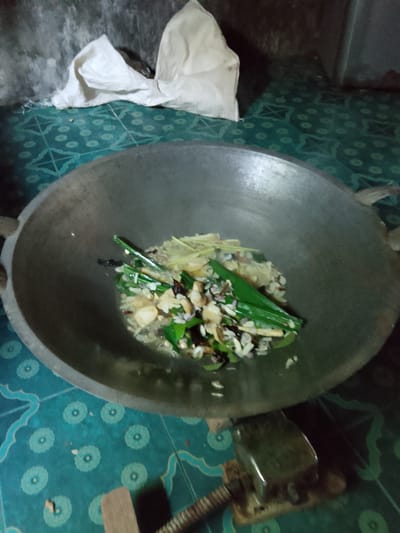 A high-angle, slightly blurry shot shows a large, dark, round wok with handles, placed on a teal-colored mat with a repeating circular pattern. Inside the wok, a mixture of ingredients is being cooked. Visible elements include sliced ginger, chopped garlic or onions, and green leafy vegetables, possibly pandan leaves, tied together. There are also some dried red chili peppers and other small, unidentifiable pieces. The background consists of a rough, grey concrete wall and a crumpled white cloth. The wok is supported by two small wooden blocks, one of which is partially visible in the foreground, with a metallic object resting on it. The overall scene suggests food preparation in a humble setting, likely in Merbopuntong, Indonesia, given the context of the location.