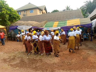 A group of women in matching outfits, including yellow head wraps, white blouses, and patterned skirts, gather outdoors, possibly for a celebration. They appear to be dancing or performing a cultural dance. Tents are set up in the background. A building can be seen in the backdrop on the left side. A man in a red t-shirt is seen standing and watching near some chairs. The ground has a reddish hue.