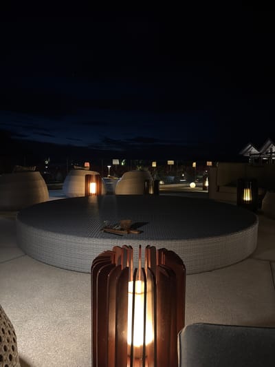The image shows an outdoor seating area at night, likely a patio or deck, in Labuan Bajo, Indonesia. To the left of the image, the dark expanse of the sea is visible. The seating area includes round, woven-style chairs and tables with lamps. In the background, there are other lights and buildings, indicating an urban or resort setting. The primary light source appears to be the lamps on the tables.