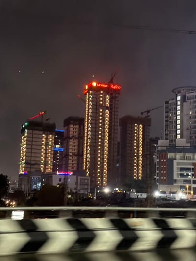 The image depicts a nighttime cityscape in Narsingi, India, focusing on a cluster of tall, illuminated buildings. The scene is characterized by the glow of lights from apartments within the structures and prominent neon signage that spells out "CAPGEEUM Skyline" in red. Several buildings appear to be under construction, indicated by cranes and scaffolding, particularly noticeable on the left side of the frame. The foreground features a blurred view of a road barrier with black and white chevron markings, suggesting the photo was taken from a moving vehicle. The sky is dark and cloudy, typical of a night sky in a metropolitan area. The overall impression is one of urban development and nighttime activity.
