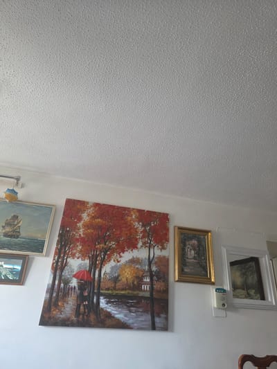 The image displays an interior room, featuring a textured white wall on the left and a smooth white wall adorned with multiple framed artworks and a wall-mounted device on the right.
The prominent feature is a large, rectangular painting depicting an autumn landscape with trees showcasing vibrant red and orange foliage alongside a body of water. Two figures are visible under a bright red umbrella, suggesting a couple embracing or walking. Below this, a partially visible painting portrays a sailing ship on a choppy sea. Above the autumn scene, a smaller, golden-framed painting shows a landscape with a path or stream. Higher up, a white-framed mirror or painting reflects a green landscape. Another small, framed picture, depicting buildings, is partially visible at the bottom.
Mounted on the smooth white wall, near the top right, is a white device with a blue and white front panel, possibly an intercom or security sensor. The text "B-TEC" is faintly visible in blue on its front. A small portion of an ornate wooden object is seen on the far right. Two small decorative ornaments, one yellow and blue, and the other light blue, hang near the bottom.
No direct activity is taking place in the room, but the artworks depict various scenes, notably an intimate autumn moment. The setting is an indoor space, likely a home, located in Cwmbrân, United Kingdom.