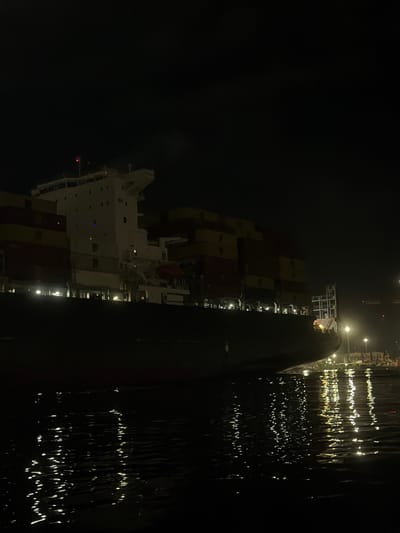 A container ship is visible at night. The ship's white superstructure, loaded with stacked containers in yellow and red, is brightly lit. Reflections shimmer on the dark water beside the vessel. Lights from a nearby shore are also reflected in the water, creating streaks of light in the darkness. The overall scene is dark, with the ship's lights providing the primary source of illumination.