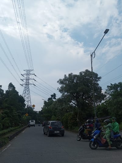 The image captures a daytime scene on a paved road lined with lush green trees, likely in a tropical or subtropical region given the foliage. The sky overhead is partly cloudy, with patches of blue visible amidst white and light gray clouds, suggesting fair weather.
Dominating the left side of the frame, a large metallic lattice power transmission tower stands tall, supporting numerous overhead power lines that stretch across the sky and diagonally down towards the left. These lines are thick and multiple, indicating a high-voltage transmission system.
Along the right side of the road, a tall, dark street light pole with a downward-angled lamp is visible. Below the lamp, there's a bent or curved metal structure attached to the pole, possibly for hanging decorations or wires, though its exact purpose isn't clear. More power lines are also visible stretching horizontally from the upper right.
The road itself is asphalt, with a faint white line marking the center. Several vehicles are present. In the midground, a dark gray hatchback car (possibly a Mitsubishi Mirage, based on its shape) is driving away from the viewer. Its license plate, partially visible, appears to be "B 2035 PVH". Further down the road, a white box truck and another car are seen, partially obscured by the car in the foreground.
On the right side of the road in the foreground, a cluster of motorcycles and riders are waiting or moving slowly. Two individuals on a blue scooter are prominent; the driver wears a dark helmet and a dark blue jacket, while the passenger, positioned behind, wears a light green or lime-colored rain poncho and a helmet, looking towards the right. Another person in a matching green rain poncho is seated further back on the same scooter. Immediately to their left, a person on a black scooter wears a dark jacket and a helmet. Other motorcycles and riders are visible behind them. The presence of rain ponchos suggests recent rain or an expectation of it, despite the partly cloudy sky.
The road curves slightly, and the surroundings are very green, with dense vegetation and trees framing both sides of the thoroughfare. In the distance on the left, near the base of the power tower, a yellow diamond-shaped road sign with a black arrow or symbol is visible, partially obscured by foliage. Another similar sign is visible further back. The scene appears to be an active, functional road in a moderately populated area, possibly an urban fringe or a suburban road.
