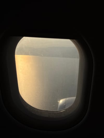 An airplane window view with a blurred landscape outside. Golden light dominates the lower portion of the window, transitioning to a hazy, brighter area above. Part of the airplane wing is faintly visible in the upper right corner. The window frame is dark and prominent, sharply contrasting with the outside light. The interior of the plane surrounding the window is dark. The window glass has visible streaks and imperfections.