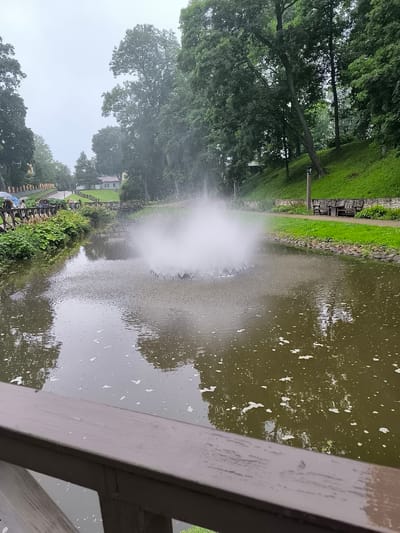 The image depicts a serene park scene in Kuldīga, Latvia, featuring a pond with a fountain in the foreground. The fountain, a prominent circular structure, sprays water upwards, creating a misty effect. Surrounding the pond is a lush green landscape with mature trees and a grassy slope. A pathway, lined with benches and lampposts, runs along the pond's edge, suggesting a public recreational area. Several people are visible walking along a wooden boardwalk in the background, some holding umbrellas, indicating overcast weather. A wooden railing in the immediate foreground frames the view, suggesting the photograph was taken from a bridge or elevated vantage point. The overall atmosphere is tranquil and inviting, characteristic of a well-maintained park.