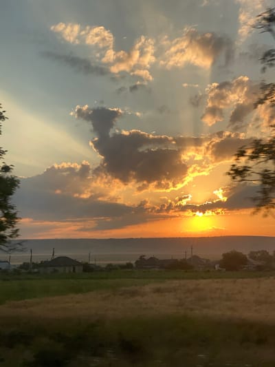 The image depicts a rural landscape at sunset, likely in or near Svetlîi, Moldova. The sky is filled with dramatic clouds, illuminated by the setting sun which casts golden rays through the clouds and creates a bright orange glow near the horizon. In the foreground, there is a field of dry grass, and further back, several houses with dark roofs are visible amidst greenery. The overall atmosphere is serene and highlights the beauty of the natural environment during sunset.
