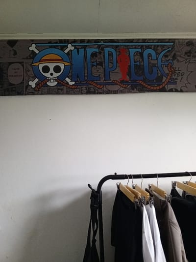 An interior scene in Al Haouafate, Morocco, depicts a white wall with a decorative banner and a clothing rack. Mounted high on the wall, near the ceiling, is a horizontal banner featuring the "One Piece" logo. The logo consists of blue "ONE PIECE" text, a skull wearing a straw hat (Jolly Roger) with crossed bones on a blue background, and integrated orange rope elements. The banner's background is a grayscale collage of manga panels, with visible text including "I HAVE MY NAKAMA!!!!", "NOTHING...", and "IT WILL NEVER EVER BE !". A red silhouette of a figure is visible within the letter 'P' of "PIECE". The majority of the wall below the banner is a plain, off-white, textured surface. In the lower right foreground, a black metal clothing rack is present. It holds several garments on wooden hangers equipped with metal clips. Visible clothing includes dark shirts, a white shirt, and a light brown garment. A black strap, potentially from a bag, hangs from the left end of the clothing rack. No individuals are visible in the scene.