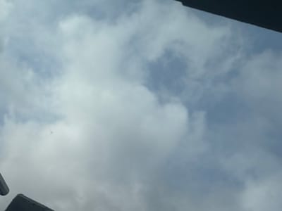 The image depicts a sky scene, asserted to be located in Germering, Germany. The overhead view shows a partially cloudy sky with formations of white and grey clouds against a backdrop of light blue sky. Dark, out-of-focus architectural elements, consistent with a roofline or building overhang, are visible at the upper right and lower left edges of the frame. A small, indistinct, light-colored airborne object is present in the upper-left quadrant of the sky. No individuals or specific ground features are discernible. The scene is illuminated by natural daylight.