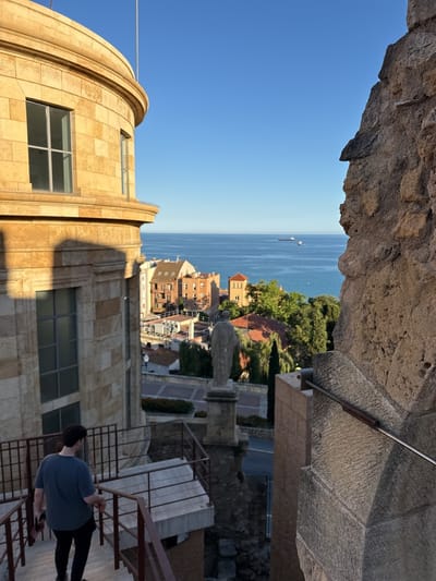 A high-angle shot in Tarragona, Spain, frames a man descending stone steps. He is clad in a short-sleeved shirt and dark pants, holding an unidentifiable object in his left hand. The steps are secured by a metal railing. To the left stands a multi-story building constructed from large, pale-yellow stone blocks. A few stories below, a statue stands centered in a green landscaped area with the city and the ocean in the background. A stone wall flanks the right side of the frame. The ocean, with at least one visible vessel, meets the clear blue sky in the background. Buildings of varying heights and architectural styles fill the space between the stairway and the water.