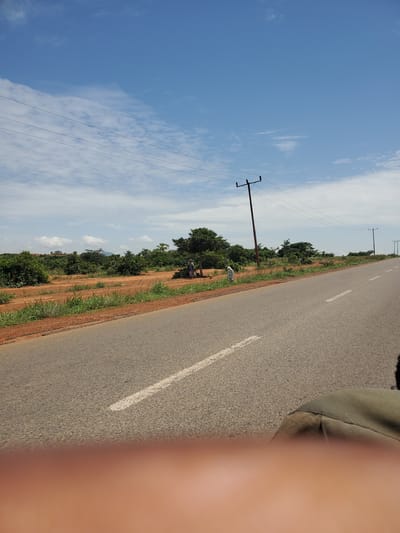 A road runs through a rural landscape in Pankshin, Nigeria. The road is paved and has a white dashed line marking the center. On the left side of the road is a dirt shoulder with sparse green grass and some shrubs. Further back are more trees and bushes. On the right side of the road, the landscape continues with similar vegetation and scattered trees. Power lines are strung across the scene, supported by wooden poles. In the distance, rolling hills can be seen under a bright blue sky with scattered white clouds. The overall scene depicts a sunny day in a rural area with evidence of electrical infrastructure.
