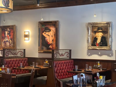The image depicts the interior of a restaurant with a Western theme, likely located in Jackson, Wyoming, United States of America, as suggested by the rustic decor and artwork. Three framed paintings adorn the wall, each with a gold ornate frame. The leftmost painting features a person on horseback in a red outfit, the central painting shows a couple in a romantic embrace, and the rightmost painting displays a woman in a cowboy hat and a black jacket.
The restaurant features red leather tufted booths and wooden tables, set with glassware and black napkins. Some tables have condiments like ketchup and yellow flowers. The lighting appears to be a mix of ambient and directed light, with overhead fixtures and wall sconces. The overall atmosphere suggests a dimly lit, cozy dining environment. There are no people visible in the image.