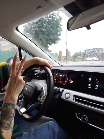 A first-person perspective shot from inside a car shows a person's hands on the steering wheel, indicating they are driving. The driver is wearing a green, yellow, and red striped sweater on their left arm, and has a tattoo of a nesting doll with flowers on their left forearm. They are also holding up two fingers in a peace sign with their left hand. The car's interior is visible, featuring a large digital dashboard with various icons and readouts. Outside the car, through the windshield, a green fence and trees can be seen, with a white car parked in the distance. The weather appears to be rainy, with water droplets visible on the windshield. The scene is likely taking place in Gol'yovo, Russia, given the context.