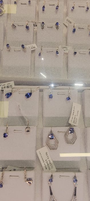 The image displays a close-up, top-down view of multiple pairs of earrings exhibited within a glass display case at a retail establishment in Mto wa Mbu, Tanzania. Thirty-three distinct pairs of earrings are visibly arranged on individual rectangular white or light gray cardboard display cards. Each earring pair features silver-toned metal settings, primarily with blue gemstones of varying cuts, including teardrop, oval, round, and raw forms, consistent with Tanzanite. Designs range from simple stud and drop earrings to more intricate geometric and elongated styles. Several display cards have small white price tags affixed, some bearing legible text such as "Artisan Galleria Ltq" and price details. Reflections on the glass surface suggest a protective barrier. The display background behind the cards is a uniform light gray.
