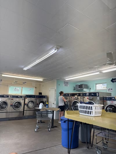 The image shows the interior of a laundromat, likely located in Ocean City, United States of America. In the background, several industrial washing machines line the wall, some of which are labeled "Dexter". A woman, wearing a gray tank top and black shorts, is standing in front of one of the machines, appearing to be loading or unloading laundry. A white plastic laundry basket filled with clothes sits on a yellow table in the foreground. To the left of the table, a blue trash can and a gray recycling bin are visible. A metal laundry cart with wheels is positioned in front of the table. The ceiling is a white drop ceiling with fluorescent lighting fixtures. The walls are painted a light blue and white. A ceiling fan is mounted near the center of the room. Several signs with instructions and warnings are posted on the walls. The floor appears to be concrete. The overall impression is of a functional and well-lit commercial laundry facility.