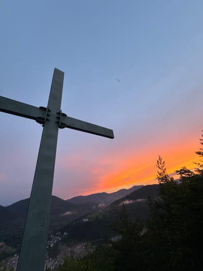 A large, dark metal cross is silhouetted against a vibrant sunset sky above a mountainous landscape. The sky transitions from a pale blue at the top to hues of orange and pink near the horizon, illuminated by the setting sun. Wispy clouds are visible, some tinged with the colors of the sunset.
The cross is positioned in the foreground, dominating the left side of the frame. It appears to be made of sturdy metal beams joined together by visible bolts and brackets. The cross is angled upwards, with the top of its vertical beam extending into the clear blue sky.
Below the cross, a valley is visible, dotted with buildings that suggest a town or village. The landscape is characterized by rolling hills and mountains covered in dark green foliage, indicating a forested area. Some patches of mist or low-lying clouds are present in the valley, adding depth to the scene. The overall impression is one of solemnity and natural beauty, with the cross serving as a focal point against the dramatic backdrop of the sunset and the natural environment. The specific location in the city of Strazha, Bulgaria, is not visually indicated by any landmarks or text within the image itself.