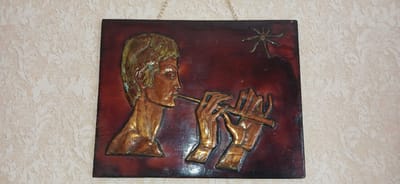 A close-up, eye-level shot shows a framed wall hanging depicting a man playing a flute. The artwork is a bas-relief with a dark red background, and the raised elements, primarily the man's profile and hands, are rendered in a metallic, copper-like finish with hints of green patina and gold. The man's head is turned in profile, facing right, with his hair appearing to be short and styled. His lips are pursed as he blows into a thin, elongated flute. His hands are positioned to play the instrument, with fingers extended and bent. Above and to the right of the man's head, a stylized, star-like object with eight points is visible. The wall hanging is suspended by a thin metal chain from the top center. The background wall is textured with a light-colored, repeating damask pattern. The lighting is somewhat harsh, casting subtle shadows on the artwork and highlighting its three-dimensional qualities. The image does not provide information about its location in Ganja City, Azerbaijan.