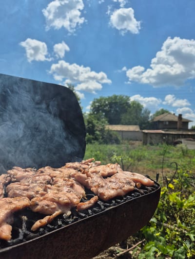 The image depicts an outdoor barbecue scene with chicken pieces being grilled on a rusty barbecue. Smoke billows from the grill, and the chicken appears seasoned and partially cooked. In the background, a rural setting is visible, with vegetation, a rustic building, and a house with a chimney. The sky is bright blue with scattered white clouds. The overall atmosphere suggests a warm, sunny day in a rural area. The location is the city of Kardam, Bulgaria.