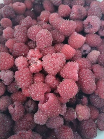 A high-angle, full-frame shot shows a pile of fresh, ripe raspberries. The berries are a vibrant pinkish-red color and are densely packed together, filling the entire frame. The lighting is natural and even, highlighting the textured surface of each raspberry. There are no people or other objects visible in the image. The scene is a close-up of a bowl or container of raspberries, with no indication of a specific location within Акбердино, Russia.