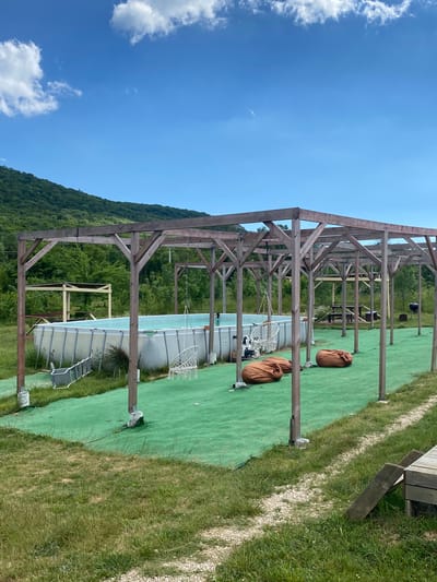 The image depicts an outdoor recreational area with a large above-ground swimming pool. The pool is surrounded by a wooden pergola structure, under which several amenities are placed. These include large, brown beanbag chairs, a white macrame hammock chair suspended from the pergola, and another similar chair on the ground next to the pool. Artificial green turf covers the ground beneath the pergola, extending around the pool area. In the background, a dense, green forest covers a hillside, under a bright blue sky with scattered white clouds. The overall scene suggests a relaxed, summer leisure setting in a rural or semi-rural environment.