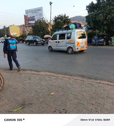 A street scene shows a young boy in a blue backpack walking on a paved sidewalk toward a crosswalk. Vehicles, including a white van and a black SUV, are present on the road. A billboard advertising "The Boulevard Schools" is visible in the background. The photo was taken with a CAMON 30S camera, using a 23mm focal length, an aperture of f/1.8, a shutter speed of 1/100s, and an ISO of 59.