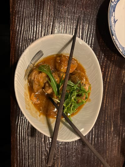 The image shows a white ceramic bowl filled with a brown, gravy-like sauce containing several pieces of meat and green leafy vegetables. The bowl is placed on a dark wooden table with noticeable wood grain patterns. Two dark wooden chopsticks are lying across the bowl, one of them fully visible, the other partially obscured by the bowl's edge. The bowl is positioned towards the bottom right corner of the frame. This location on the island of Naoshima, Japan, is characterized by a variety of contemporary art installations, including the Benesse House Museum and the Chichu Art Museum, and the image was taken from a restaurant inside one of these facilities.