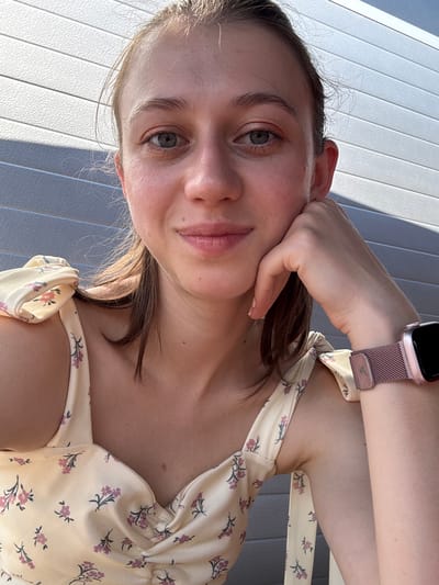 A young woman with fair skin and light brown hair tied up is looking at the camera. She has blue eyes and is resting her chin on her hand. She is wearing a light yellow dress with a floral pattern. On her left wrist, she is wearing a rose gold smartwatch. The background is a gray wall made of horizontal planks. The lighting suggests it is daytime and the sun is casting shadows on the wall. The image appears to have been taken outdoors. There is no discernible indication of the location in Obernburg am Main, Germany.