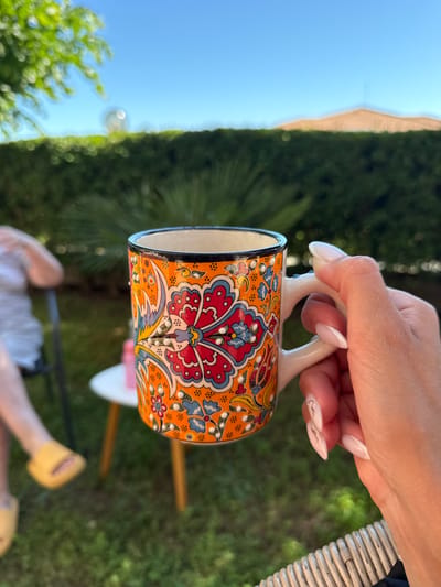 A close-up shot captures a hand holding an ornately decorated orange mug with floral patterns in red, blue, and yellow. The mug has a white handle and a black rim. The background is a soft-focus, sunny outdoor scene, featuring lush green foliage and a clear blue sky. To the left, a person's legs are visible, wearing yellow slides and sitting on a chair. In the mid-ground, a small white table with wooden legs is present. The scene appears to be set in the city of Kosharitsa, Bulgaria, given the architecture visible in the blurred background which suggests a Mediterranean climate. The overall impression is one of relaxed outdoor leisure.