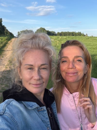 Two women are captured in a close-up, rotated outdoor portrait, likely a selfie, with their heads close together. The image is rotated 90 degrees counter-clockwise, making the sky appear to the left and the ground to the right.
The woman in the upper portion of the frame has long, straight, warm-toned blonde hair and is wearing a light pink zippered jacket. She has a subtle smile and is looking towards the camera, with light-colored eyes. Her hand is visible near her chest, seemingly resting on her jacket. She wears large hoop earrings.
Below her, the second woman has shorter, curly, cool-toned blonde hair. She is wearing a black top layered under a denim jacket. She also smiles faintly and looks directly at the camera with light-colored eyes.
The setting appears to be a rural or natural area, possibly in Норд Снт, Russia. In the background, there is a vibrant green field, transitioning to a line of trees in the distance, all under a clear blue sky with a few wispy clouds. The lighting suggests late afternoon or early evening, with a soft golden glow on their faces and hair. Both women appear relaxed and happy, sharing a friendly moment in the open air. There is no visible text in the image.