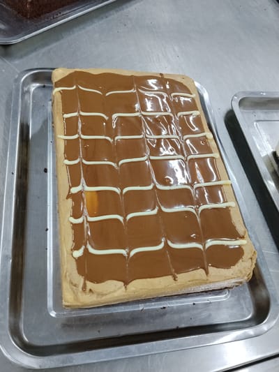 A rectangular cake is presented in a silver tray on a stainless steel surface. The cake is topped with a smooth, dark brown frosting that has a glossy sheen. Over this, thin white lines are drizzled in a decorative pattern, creating a grid-like appearance with curved lines within each square. The cake itself appears to have a light brown sponge base. In the background, another silver tray with a dark-colored dessert is partially visible. The overall environment suggests a commercial kitchen or bakery setting.