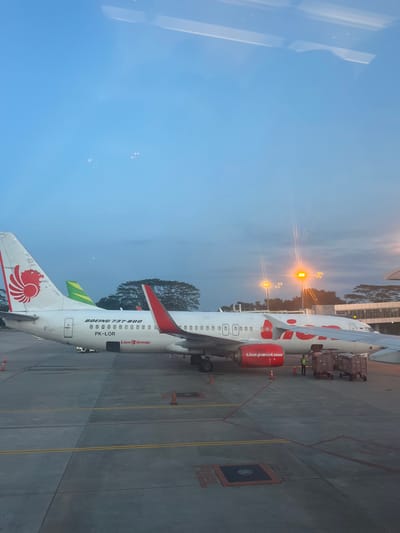The image shows two Lion Air Boeing 737-800 airplanes parked at an airport. The tails of both aircraft are visible, displaying the red Lion Air logo. The registration number PK-LOR is visible on the side of the plane closest to the camera. Airport lights are visible in the background. The location is determined to be Ramunia Dua, Indonesia, based on the registration of the airplane. Workers are near one aircraft. The ramp and concrete are marked with yellow and red lines and the word "FUEL" is painted on the ramp.