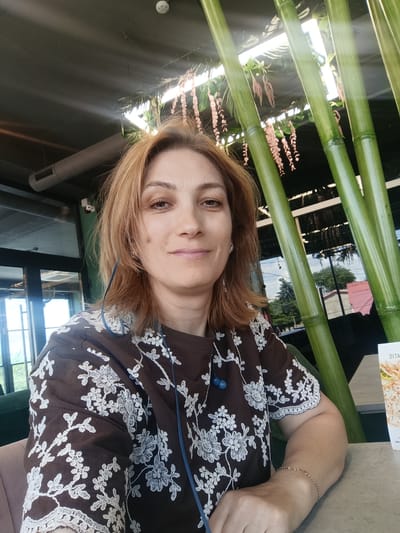 A medium shot captures a woman with auburn hair and light skin, positioned slightly to the left of the frame, in what appears to be an indoor establishment. She is looking directly at the camera with a gentle smile, her head tilted slightly. She wears a brown shirt adorned with a white floral embroidery pattern. Blue earbuds are visible, with the cord draped over her shirt. Her right arm rests on a table, on which a menu is partially visible, displaying the text "SIT A".
Behind her, several large, vibrant green bamboo stalks rise vertically, partially obscuring the background. The environment suggests an indoor seating area, possibly a restaurant or cafe, with a dark ceiling and ambient lighting. Reflections in the glass doors behind the bamboo hint at an outdoor scene with trees and buildings under a bright sky. The overall lighting is natural and diffused, creating a warm and inviting atmosphere. The woman is wearing a thin bracelet on her right wrist. The exact location within the city of Florești, Moldova, is not visually ascertainable from the image.