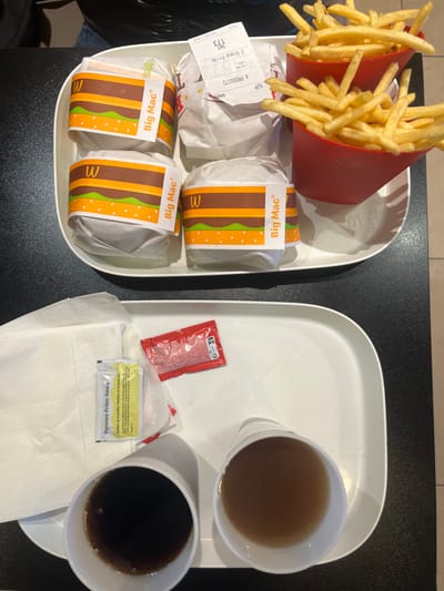 The image displays two white rectangular fast-food trays on a dark, possibly black, tabletop, consistent with an interior setting of a McDonald's restaurant in Fontenay-sous-Bois, France.

The upper tray contains four individually wrapped items. Three of these items are clearly identified by their paper bands as "Big Mac" sandwiches. The fourth wrapped item is visually similar. Two red, ribbed plastic cups, each filled with golden-brown French fries, are also present on this tray. A white paper receipt, partially visible on one of the wrapped items, indicates "2 Grand Frit" (two large fries) and "4 PRODUITS" (four products).

The lower tray holds two white paper cups; one contains a dark brown carbonated beverage, and the other contains a lighter brown liquid. A stack of white paper napkins is positioned on the left side of this tray. Next to the napkins are two small foil packets of condiments: one yellow packet labeled "Pommes-Frites-Sauce" (French for fries sauce) and one red packet, likely containing ketchup.

A partial view of an individual's knee or lap, clad in dark fabric, is visible in the upper left corner of the frame, indicating the presence of a person seated at the table.
