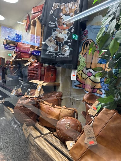 The image shows a display of leather bags and t-shirts in a shop window. The display features various styles and colors of handbags, as well as t-shirts with graphic prints. A plant is visible on the right side of the frame. The text on some of the tags indicates that the items are from "GLAMOUR" and some prices are visible, such as "€1.995" and "€99.95". Some digital information with train schedules is reflected in the glass, suggesting the shop is located in a station or a high-traffic area. One of the reflections shows "Duisburg Hbf", indicating the shop is located in Duisburg Hauptbahnhof (central station), Germany.
