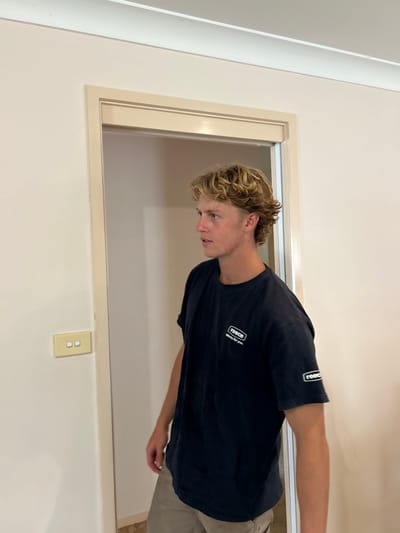 An interior scene in Thirroul, Australia, depicts a young Caucasian male standing near a doorway. The male, approximately late teens to early twenties, has fair skin, short blonde curly hair styled in a mullet, and a neutral expression as he looks towards the left of the frame. He is wearing a dark blue short-sleeved t-shirt with a white "reece Waiting for you" logo on the left chest and a similar logo on the left sleeve, paired with light brown trousers. He stands with his left arm slightly bent, his right arm partially obscured. To his left, mounted on an off-white wall, is a cream-colored double light switch. The doorway features a light cream-colored architrave. The walls are painted off-white, and a white cornice is visible at the ceiling line. The flooring visible through the doorway is light-colored.