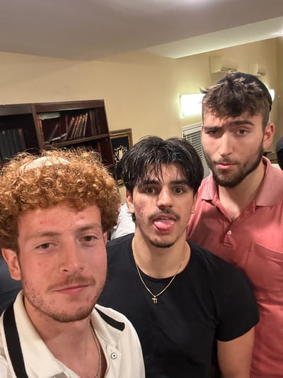 A photograph captures three young adult males posing indoors. The leftmost individual, in the foreground, has curly reddish-brown hair, a light beard, and an apparent yarmulke beneath his hair, wearing a light-colored collared shirt with dark piping; he looks directly at the camera. The central figure has dark hair, a light mustache, wears a black t-shirt with a gold 'Chai' pendant, and is sticking his tongue out towards the camera. The third individual, on the right, wears a dark yarmulke on his head and a red polo shirt, with a beard, and holds a serious expression while looking forward. The background features a dark wooden bookshelf filled with books, an ornate framed artwork, and a cream-colored wall with a rectangular light fixture. The scene is located in Bergenfield, United States, consistent with a communal or institutional setting.