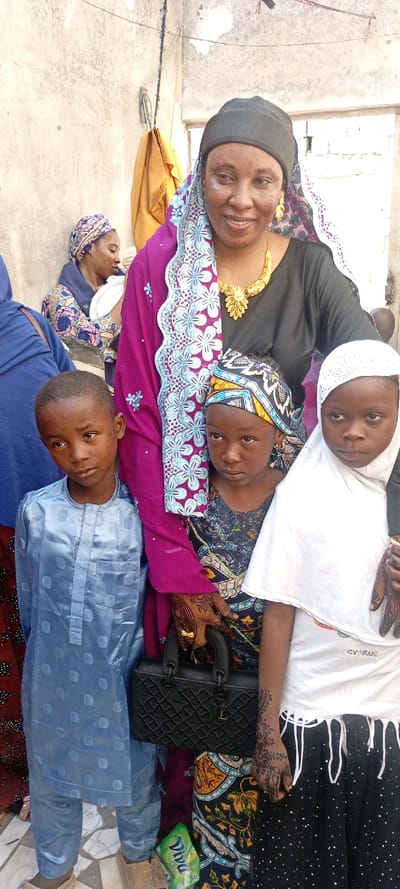 An outdoor, eye-level shot in Dan Maliki, Nigeria, shows five individuals in the foreground and midground, with two additional figures partially visible in the background, against a rough, light-colored concrete wall.
In the center, a woman in a black head covering, black top, and a magenta-pink outer garment with light blue floral embroidery smiles towards the camera. She wears prominent gold-colored earrings and a matching necklace. Her right hand, decorated with henna, holds a black, textured handbag.
To her left, a young boy in a light blue traditional tunic and trousers stands with a neutral expression. To her right, a young girl with a patterned headscarf and dark patterned dress stands behind the boy, also with a neutral expression. Adjacent to this girl, on the far right, another young girl wears a white perforated hijab, a white t-shirt with "CYPRUS" text, and a black polka-dotted skirt with a fringed hem. Her right hand also displays henna.
In the left midground, partially obscured, a woman in a blue garment and head covering is visible. Further left in the background, another woman is seen in profile, wearing a patterned headscarf and dress, appearing to hold a white bundle.
The background consists of a worn, light-colored wall with an orange fabric piece hanging on it, and a doorway opening on the right. A green "Diva Plus" package lies on the ground in the lower-right foreground.