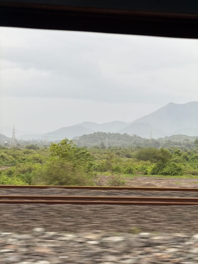The image depicts a landscape seen through a window, likely from a moving vehicle. In the foreground, two parallel train tracks are visible, with the ballast beneath them blurred due to motion. Beyond the tracks, a dense expanse of green foliage covers the terrain. In the mid-ground, rolling hills are dotted with trees and vegetation, and a prominent electricity pylon stands on the left side. Further in the distance, a series of hazy, layered mountains fade into the overcast sky. The overall impression is that of a rural or semi-rural environment under a cloudy or misty day. The image is likely taken in Vasai-Virar, India, a region known for its green cover and mountainous terrain.