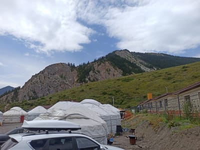 The image shows a campground with yurts and buildings in the city of Kurmeti, Kazakhstan. The scene is dominated by a large mountain in the background, covered in green grass and trees. In the foreground, there are several white yurts, some of which are covered with tarps. A white SUV with a roof box is parked in front of the yurts. There is also a building with a red roof and a black fence, and several chairs and a barbecue grill are scattered around the campsite. The sky is cloudy with some patches of blue. The overall impression is one of a remote and rustic camping location in a mountainous region.