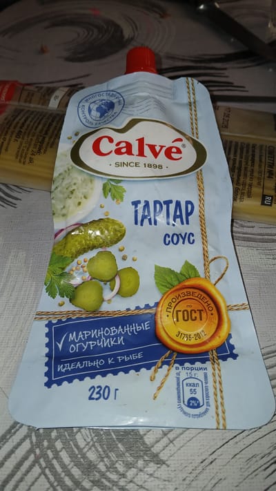 A close-up, eye-level shot captures a squeeze pouch of Calvé Tartar Sauce, predominantly white with blue accents, against a textured, grey and white surface. The pouch features prominent branding, including the "Calvé" logo and "SINCE 1898." Below this, "TARTAR SOUS" is written in large, blue, sans-serif font. Decorative elements include illustrations of pickles, onions, herbs, and a seal indicating GOST certification. Nutritional information is visible in a circular inset, showing "55 kcal" per 15g serving, which represents 2% of a recommended daily intake. The packaging also states "MARINATED CUCUMBERS" and "IDEAL FOR FISH" in Russian. The background suggests other food packaging, possibly pasta. There is no visible indication of the city of Kolyvan, Russia, in this image.
