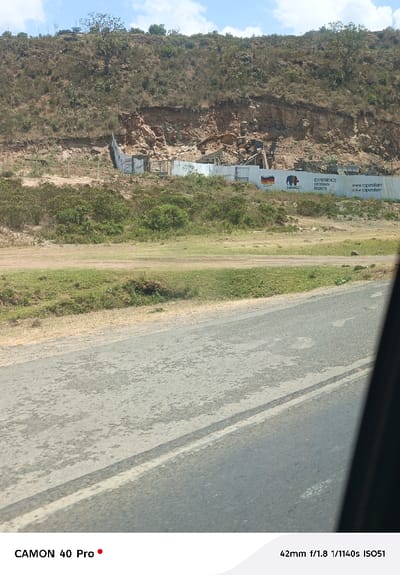 The image depicts an outdoor scene in Gilgil ward, Kenya, viewed from a vehicle. A two-lane asphalt road with a faded white centerline occupies the lower foreground, partially obscured on the right by a dark vehicle door frame. Beyond the road is a dirt and grassy shoulder with scattered low green bushes. In the midground, a construction site is visible, enclosed by a white corrugated metal fence. This fence features advertising for "Caparol" German paints, displaying a German flag and an elephant logo, along with text "EXPERIENCE GERMAN PAINTS" and a website address. Behind the fence, several pieces of heavy construction machinery, including excavators and loaders, are visible amidst excavated earth and rubble. The background is dominated by a dry, rocky hillside or cliff face with exposed reddish-brown soil and sparse, arid vegetation including scrub and occasional trees. The sky is light blue with scattered white clouds. No people or explicit actions are discernible.