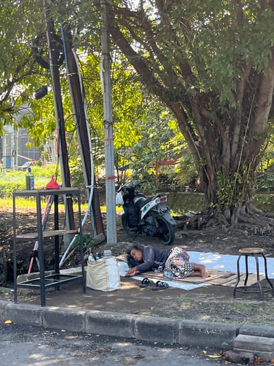 In Pagerwojo, Indonesia, a person sleeps on a mat on the ground next to a large tree with exposed roots. A motorcycle is parked nearby. To the left of the sleeping person, a tan bag and a small metal shelf unit with various items on it are visible. The scene is outdoors, with sunlight filtering through the leaves of the tree, casting shadows on the ground. A small wooden stool is positioned to the right of the sleeping person. The ground is a mix of dirt and paved surfaces, with a concrete curb running along the edge of the road. Electrical poles and wires are present in the background, along with some distant structures and greenery.